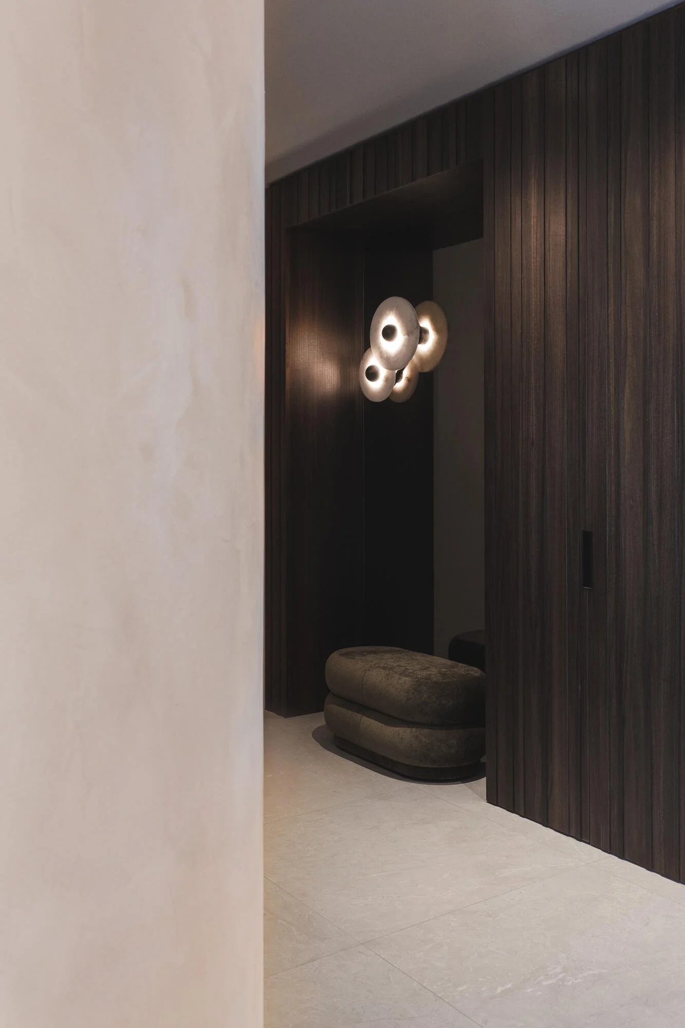 A modern interior space with a dark wood wall, a sectional cushioned seat, and a sculptural ceiling light fixture with three concentric rings. Part of a light-colored wall is visible in the foreground.