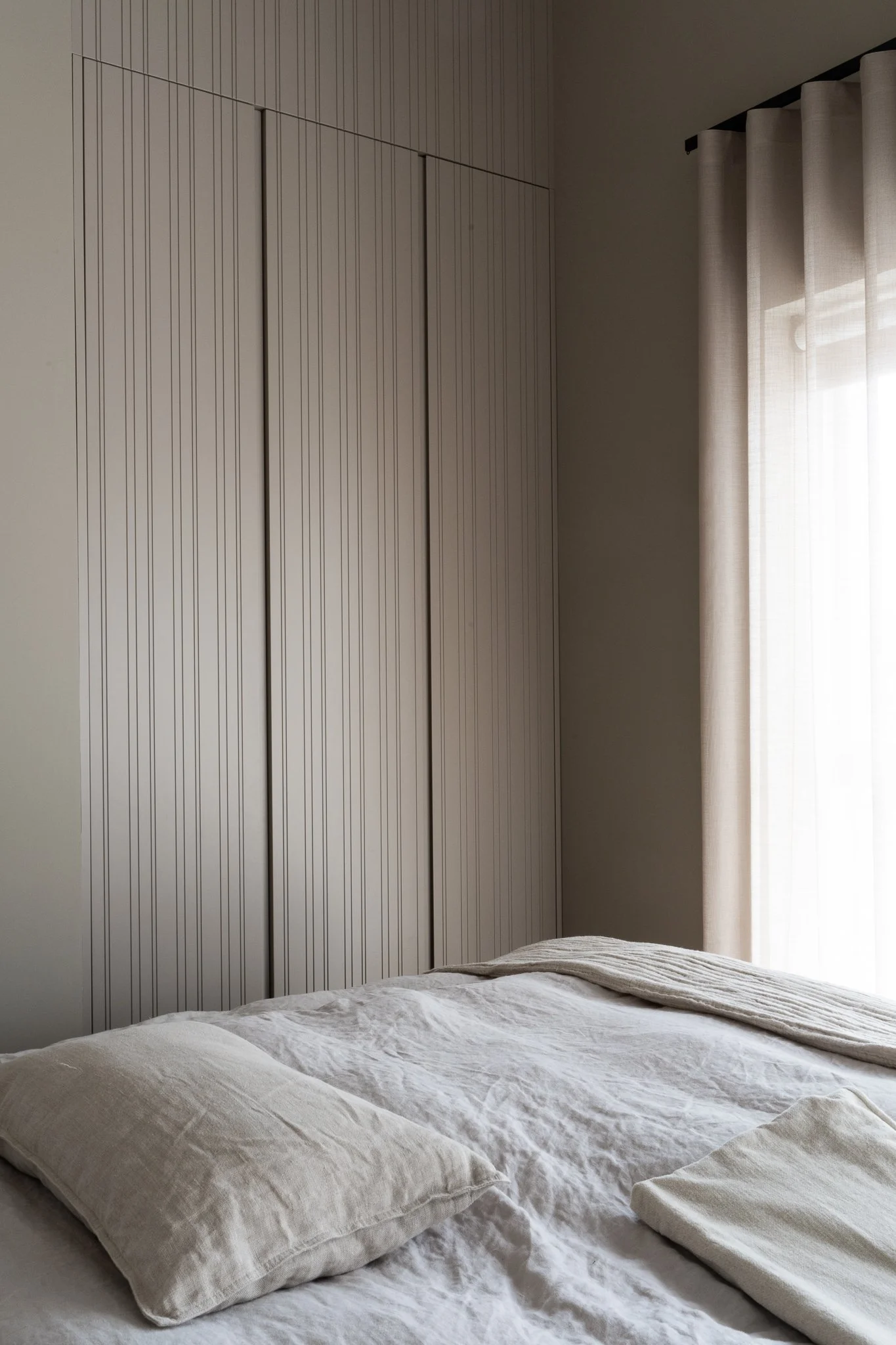 A neatly made bed with white linen, large pillows, and a throw blanket in a bright room with beige curtains and a textured wall.