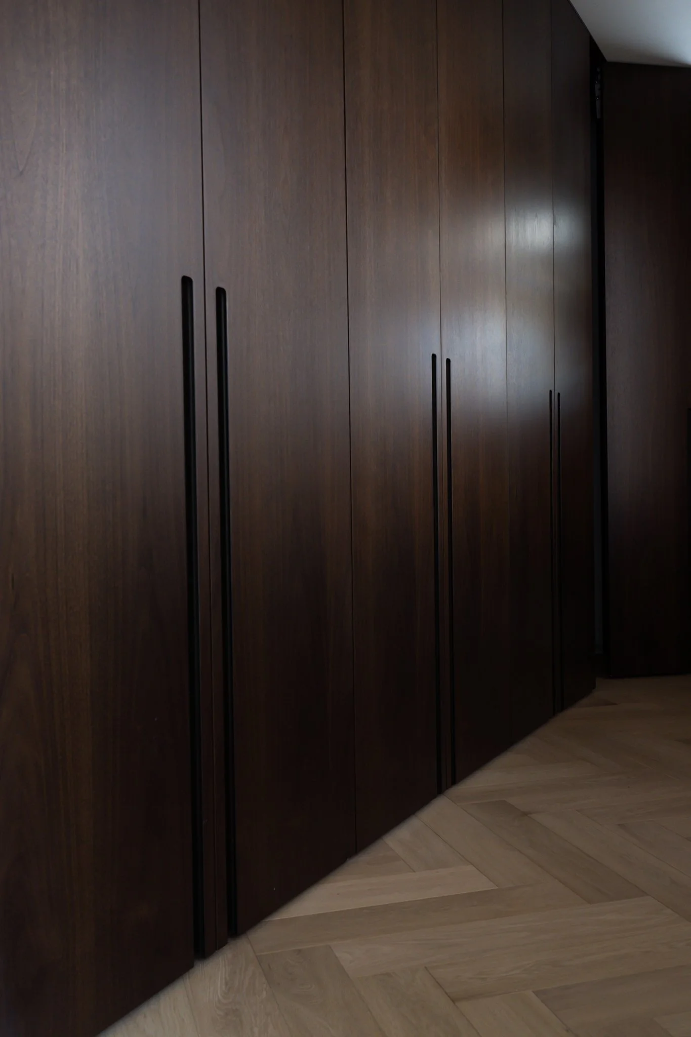 A dark wooden wardrobe with three doors and black handles, situated next to a corner wall and light-colored wooden floor.