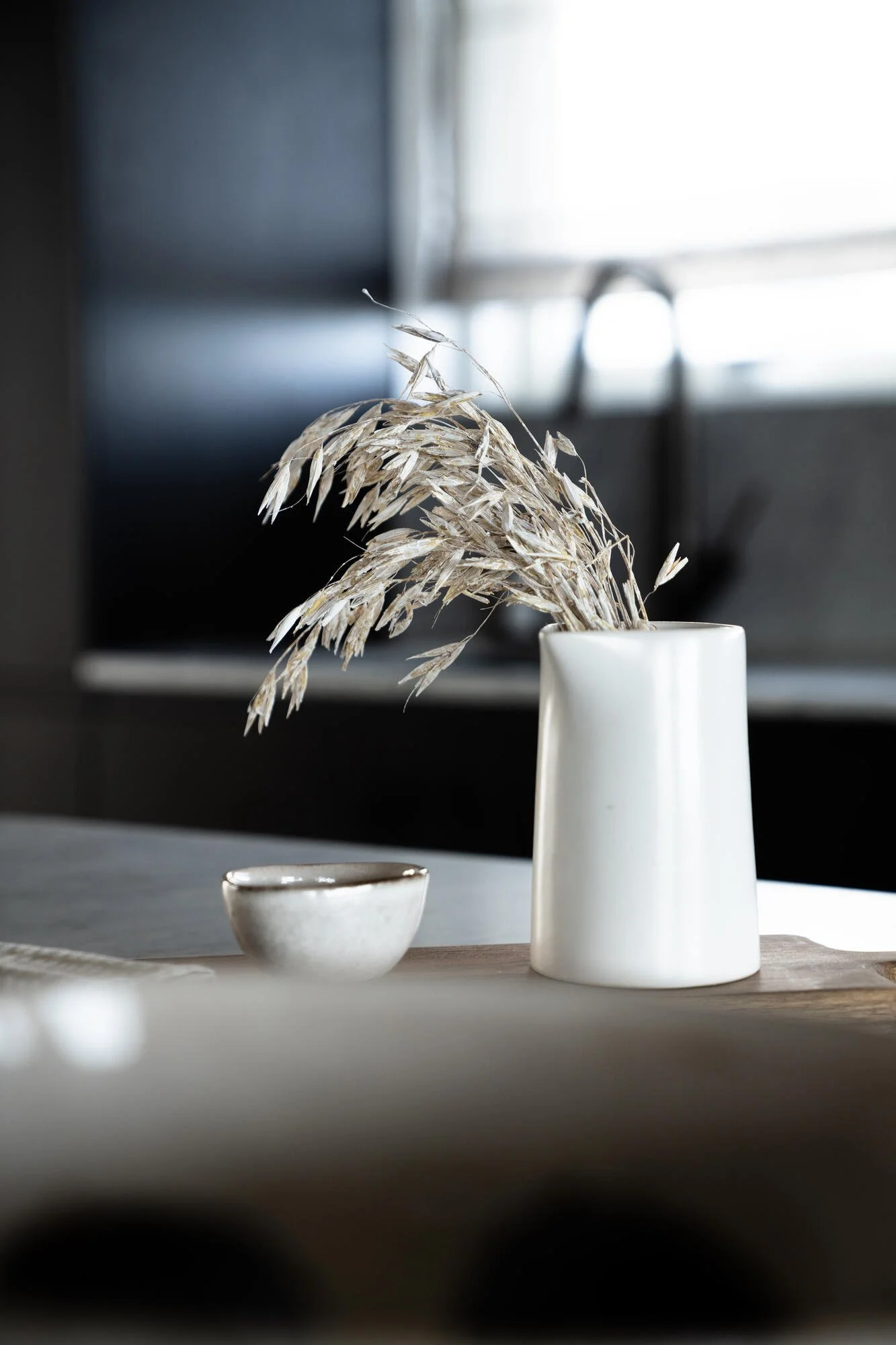 Minimalist white ceramic vase with dried grasses, a small ceramic bowl, and a light-colored wooden board placed on a table, in front of a blurred background with natural light coming through a window.