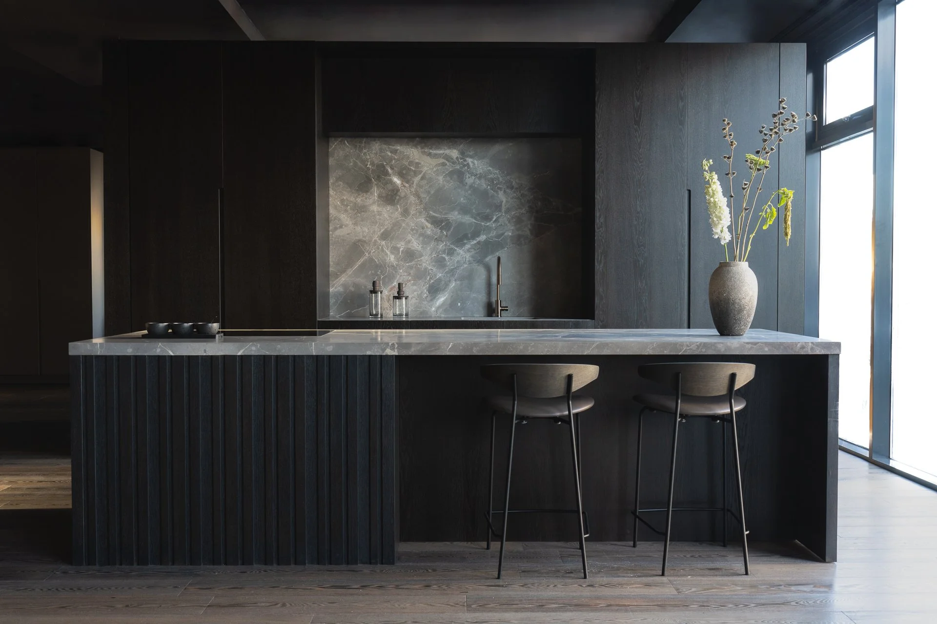 Modern kitchen with black cabinetry, marble countertop, and a vase of tall white and green flowers by a large window.