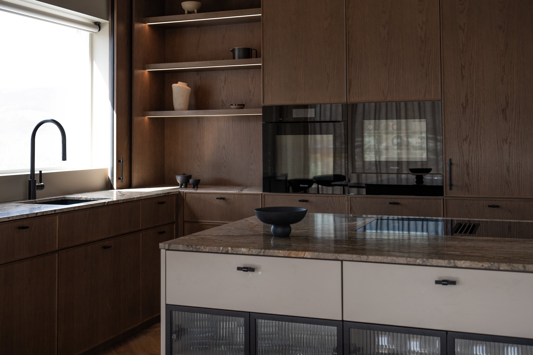 A modern kitchen featuring wooden cabinetry, a marble countertop, a black sink with a high-arc faucet, and built-in appliances including an oven. Open shelves hold decorative vases and bowls, with a window allowing natural light to illuminate the space.