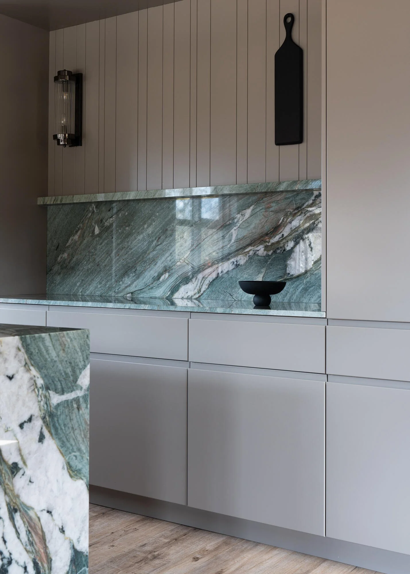 Modern kitchen with beige wooden panel wall, green marble countertop, white cabinets, black wall decor, and a black bowl on the counter.