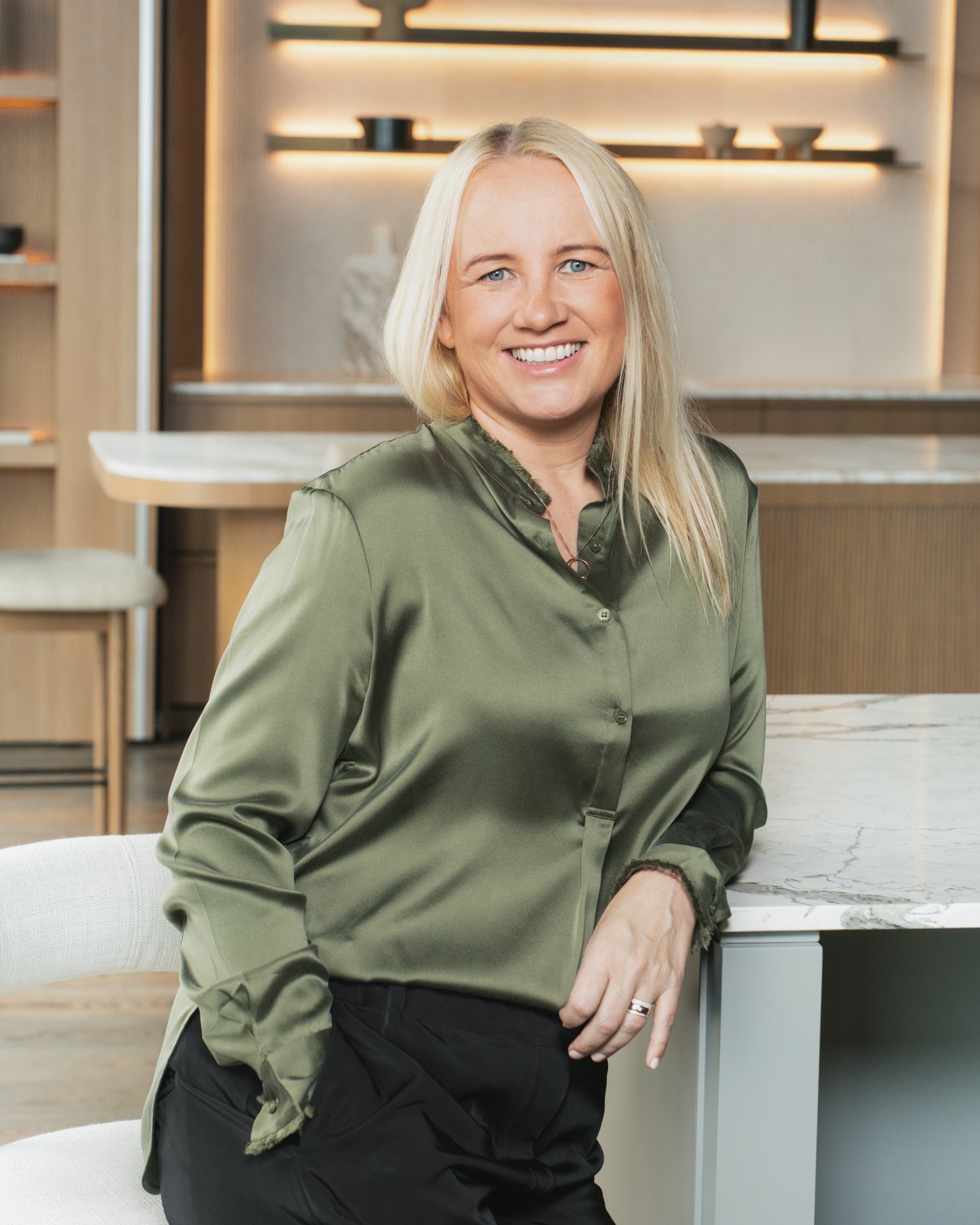 A smiling woman with blonde hair wearing a green satin blouse and black pants, sitting at a marble kitchen counter in a modern kitchen with open shelving and decorative objects in the background.