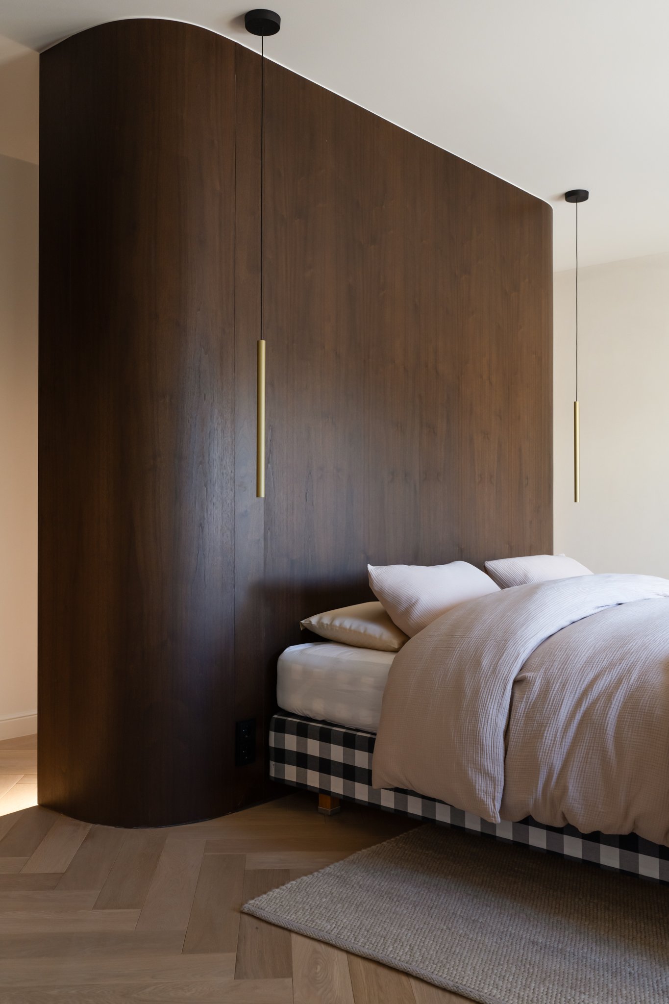 A bedroom with a dark wooden curved headboard, two gold pendant lights hanging from the ceiling, a bed with white and beige bedding, placed on a checkered bed base, and a light-colored rug on a wooden floor.