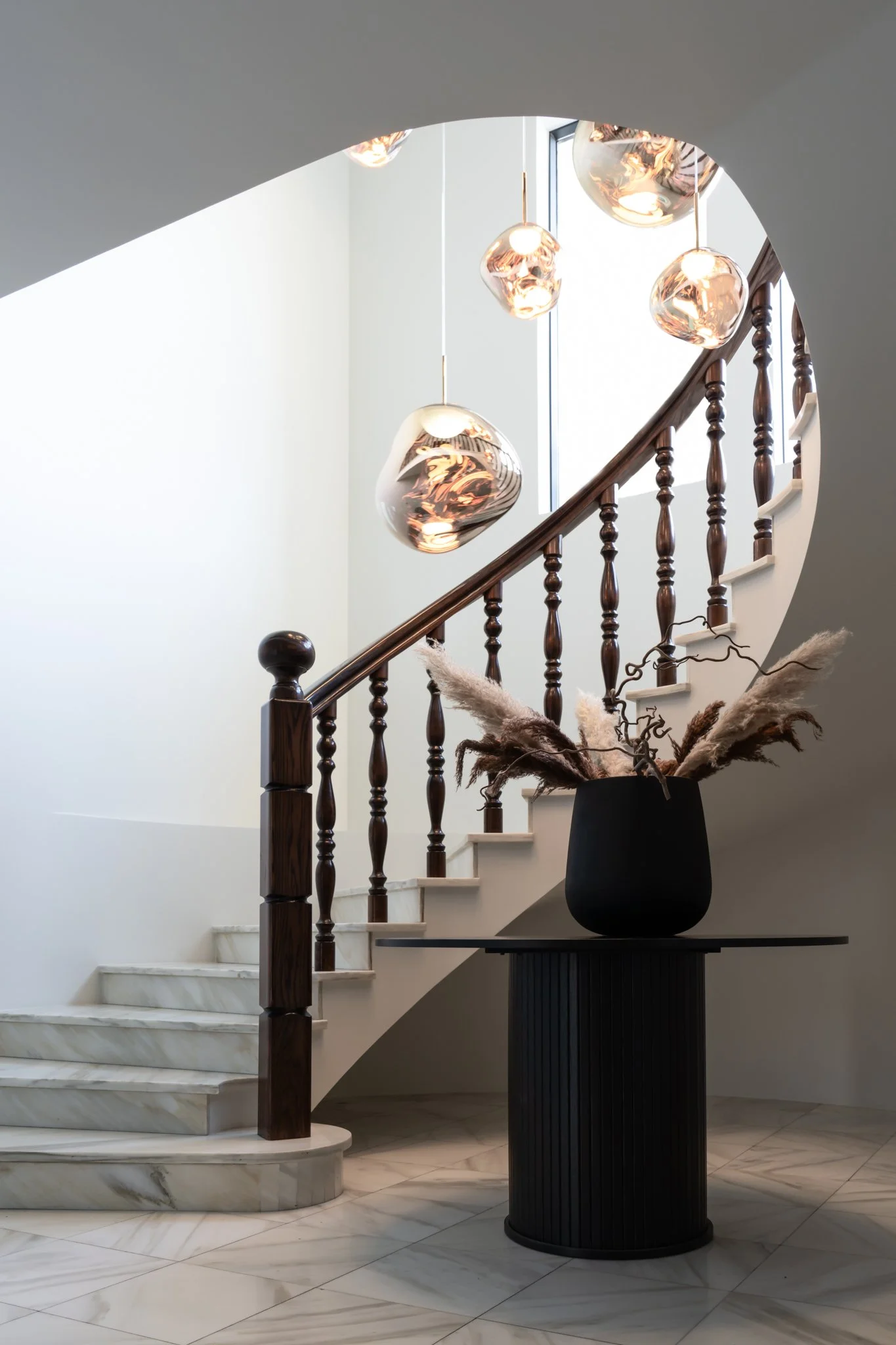 An interior staircase with white marble steps, wooden handrail, and modern hanging pendant lights, with a large black vase holding dried ornamental grasses nearby.