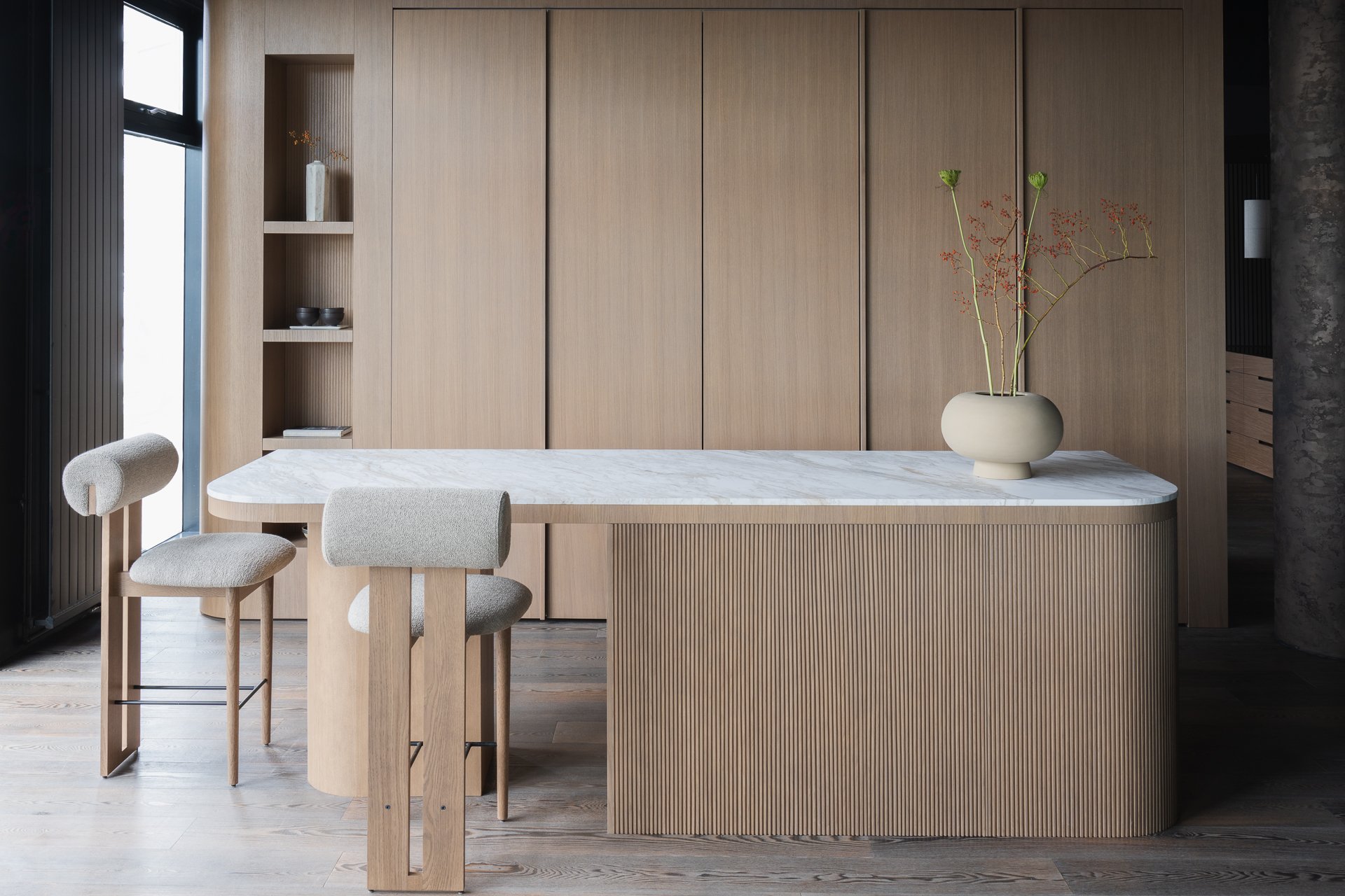 Modern dining area with a light wood table, beige upholstered chairs, and a large light wood cabinet wall. There's a white marble countertop and a minimalist beige vase with a branch of red and green decorative flowers.