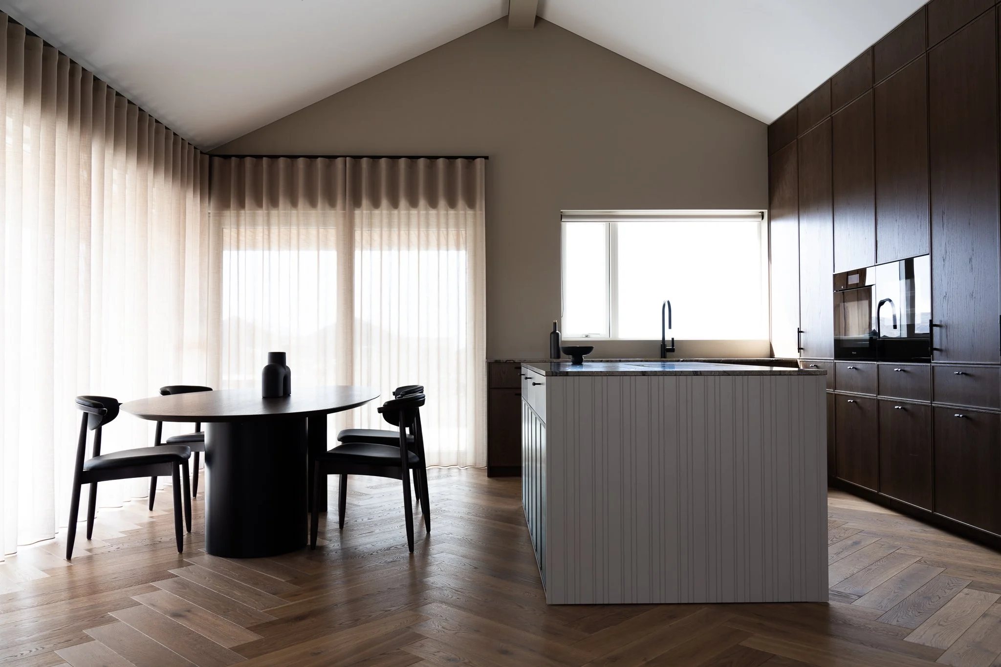 Modern kitchen with dark wood cabinets, white walls, a window above the sink, a small dining table with black chairs, and wooden flooring.