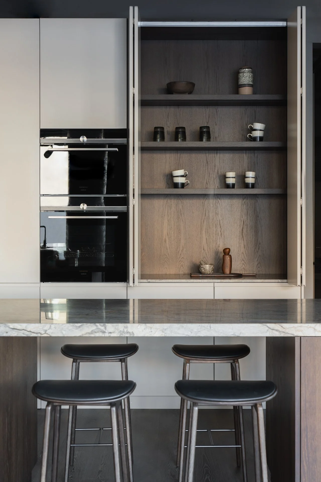 Modern kitchen with a marble island and black bar stools, open cabinets with dishes, and a built-in oven.