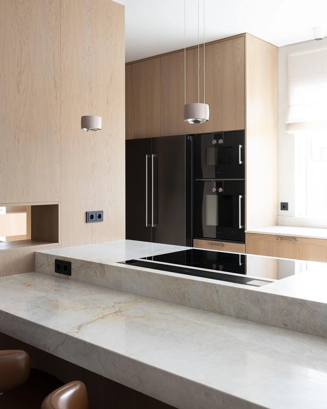 Modern kitchen with light wood cabinetry, black built-in oven and refrigerator, white marble countertops, and hanging pendant lights.