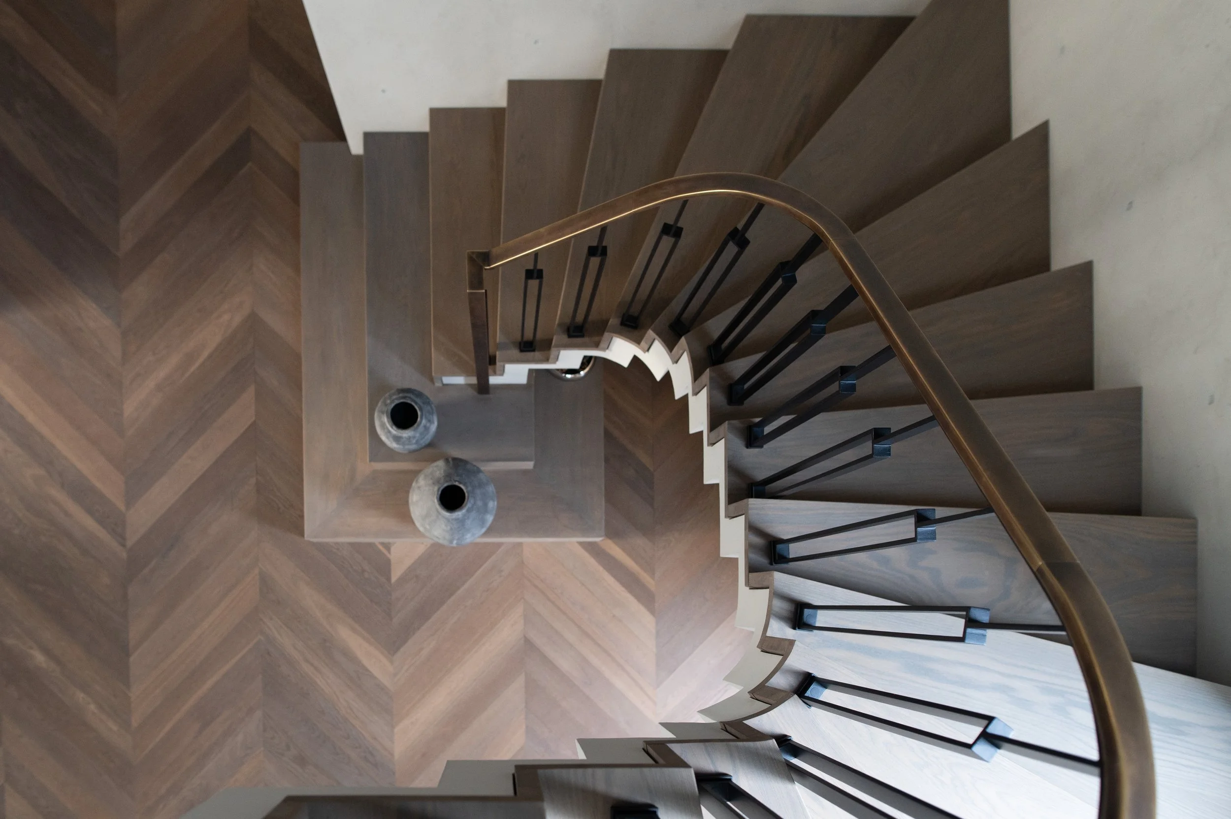 A view down a spiral staircase with dark wooden steps, black rails, and a gold-colored handrail. At the bottom, two round marble tables are visible on a herringbone wood floor.