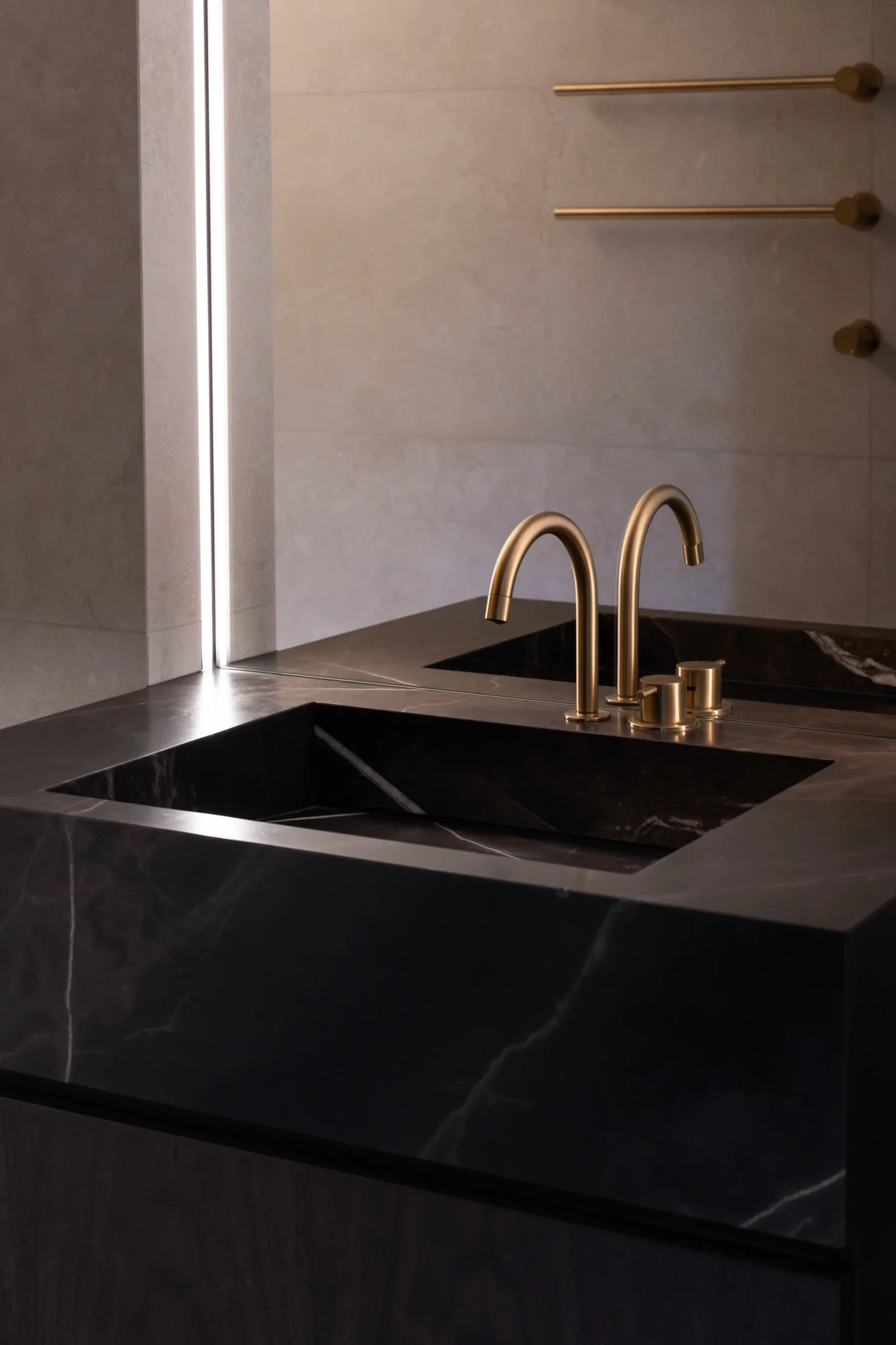 A bathroom sink with two gold-colored faucets on a black marble countertop, with a beige tiled wall behind.