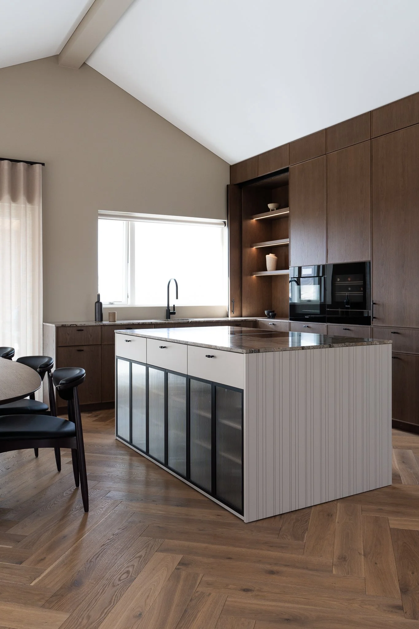 Modern kitchen with wooden cabinets, a marble island, black chairs, and a large window letting in natural light.