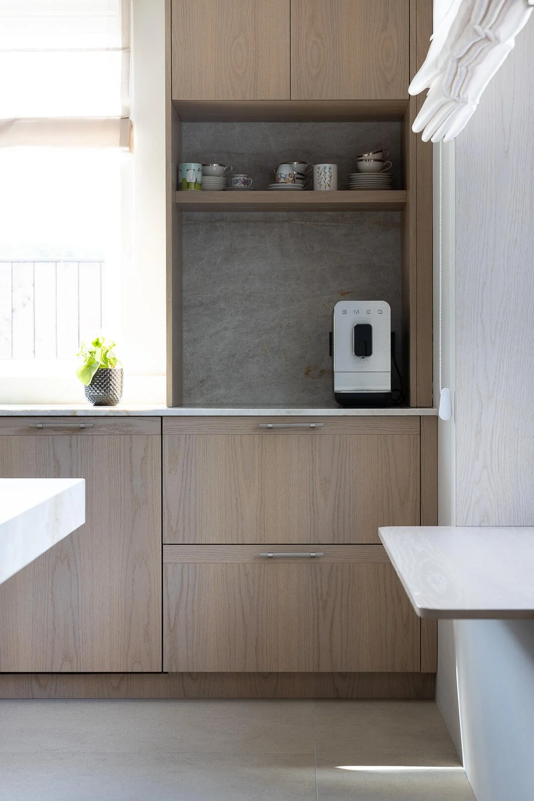 Kitchen with light wood cabinets, a window with a plant, a coffee machine, and shelves with teacups and plates.