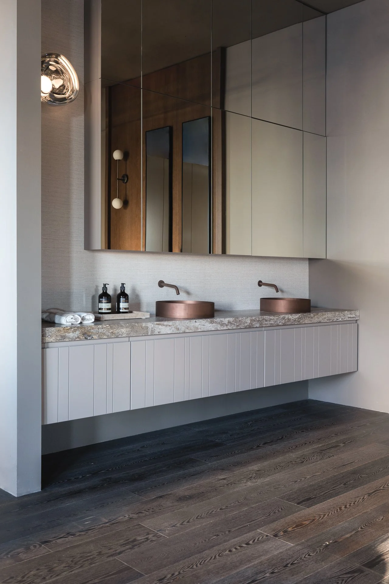 Modern bathroom with floating vanity, two copper sinks, large mirror, dark wood floors, and minimalistic decor.