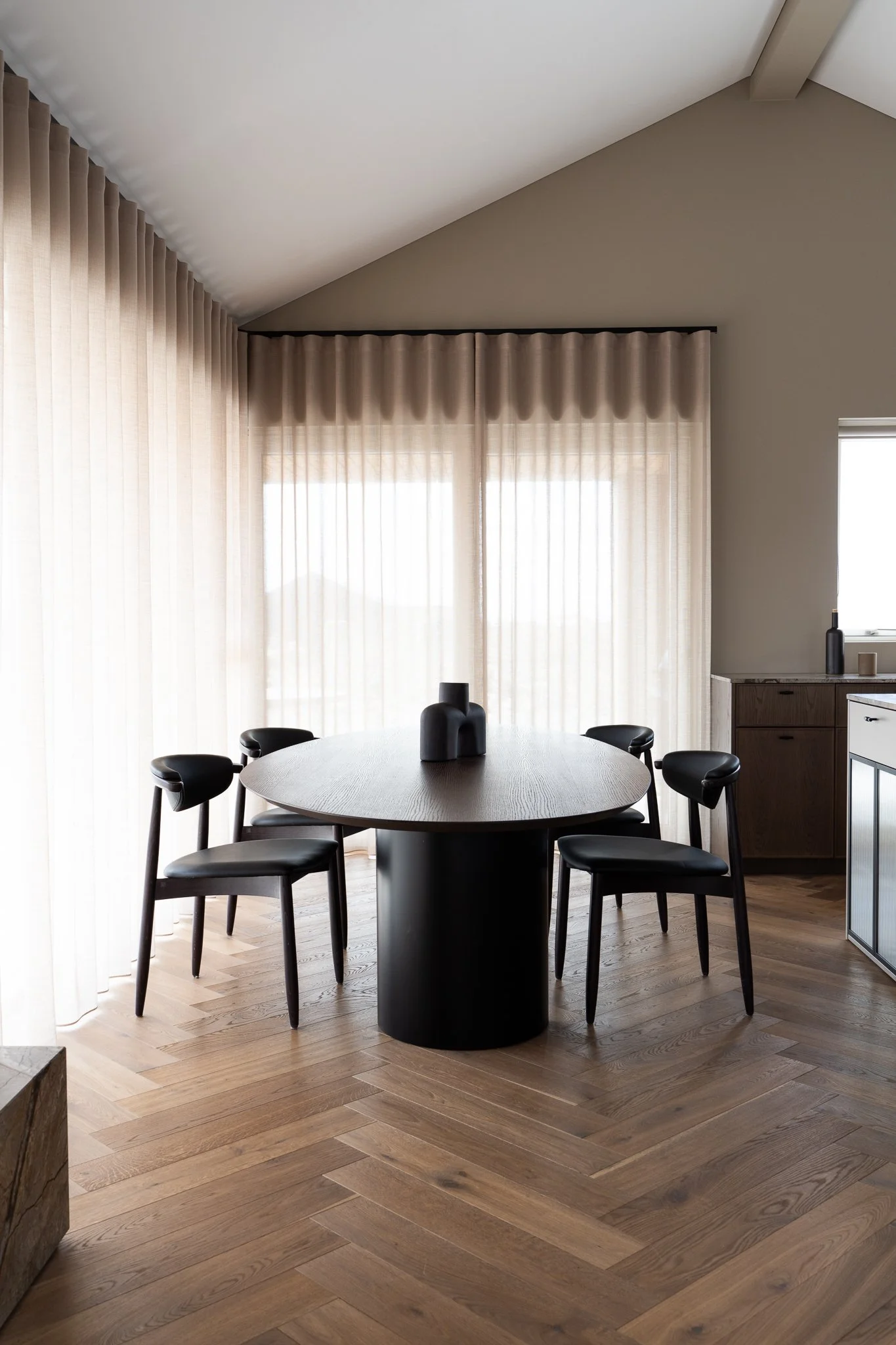Modern dining room with a round black table and six black chairs. There are beige curtains covering large windows, and a small wooden cabinet against a beige wall. The room has a sloped ceiling and wooden flooring.