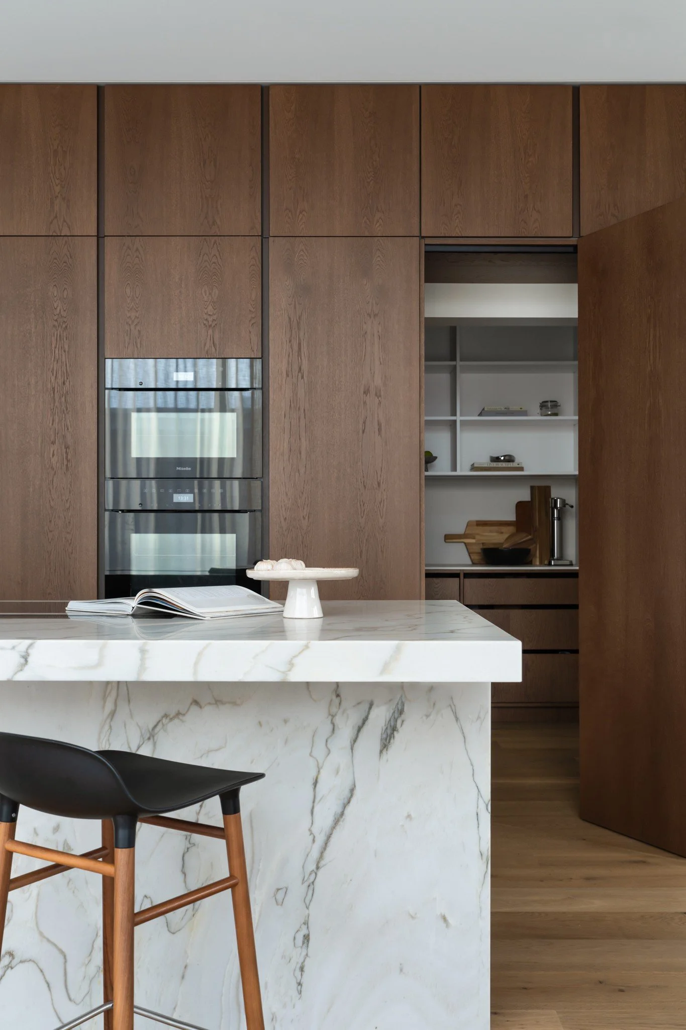 Modern kitchen with a white marble island, wooden cabinets, built-in oven, open shelves, and a black chair with wooden legs.