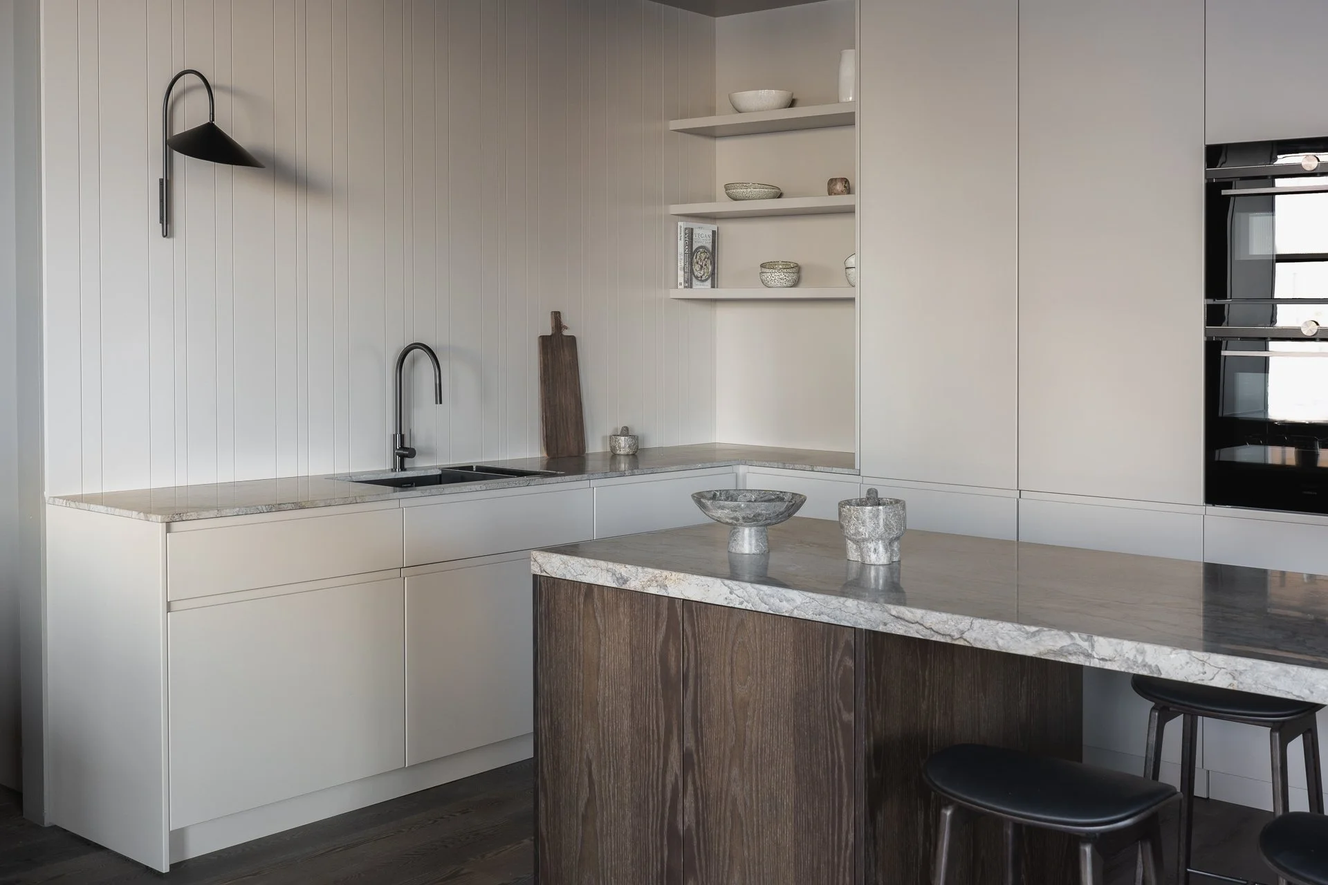 Modern kitchen with white cabinets, a gray marble countertop, a small sink, and open shelves with bowls and books. There are black chairs at a breakfast bar and a dark wooden kitchen island with a marble top.