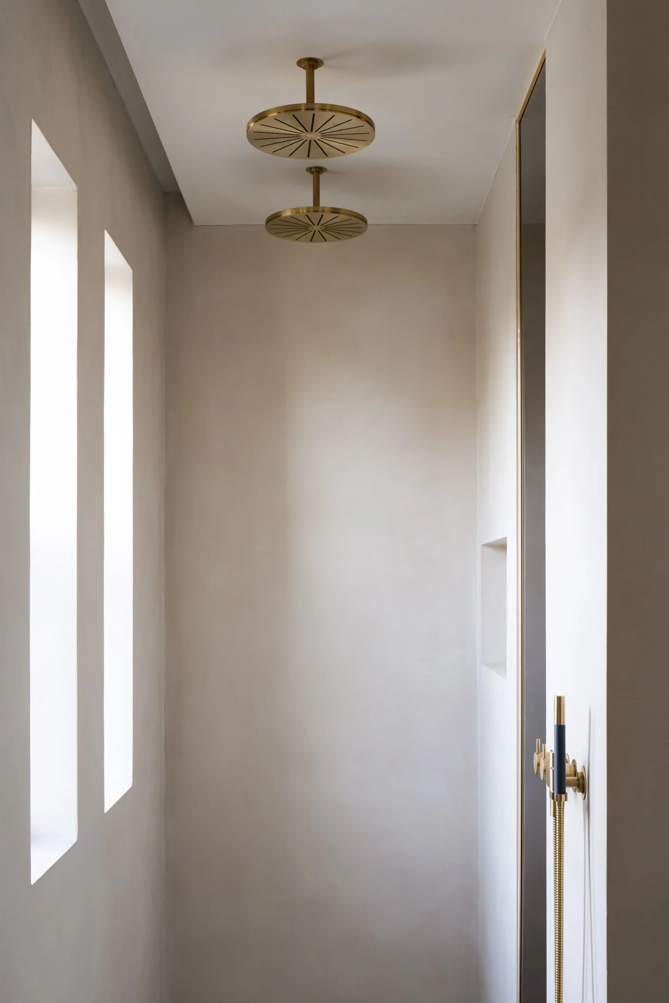 A narrow, minimalist hallway with white walls, three vertical windows on the left, and a modern gold and black lighting fixture on the ceiling.