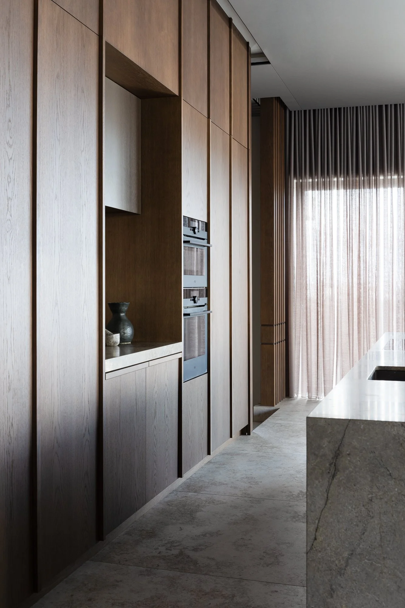 Modern kitchen interior with wooden cabinets, a granite countertop, and sheer curtains covering large windows.
