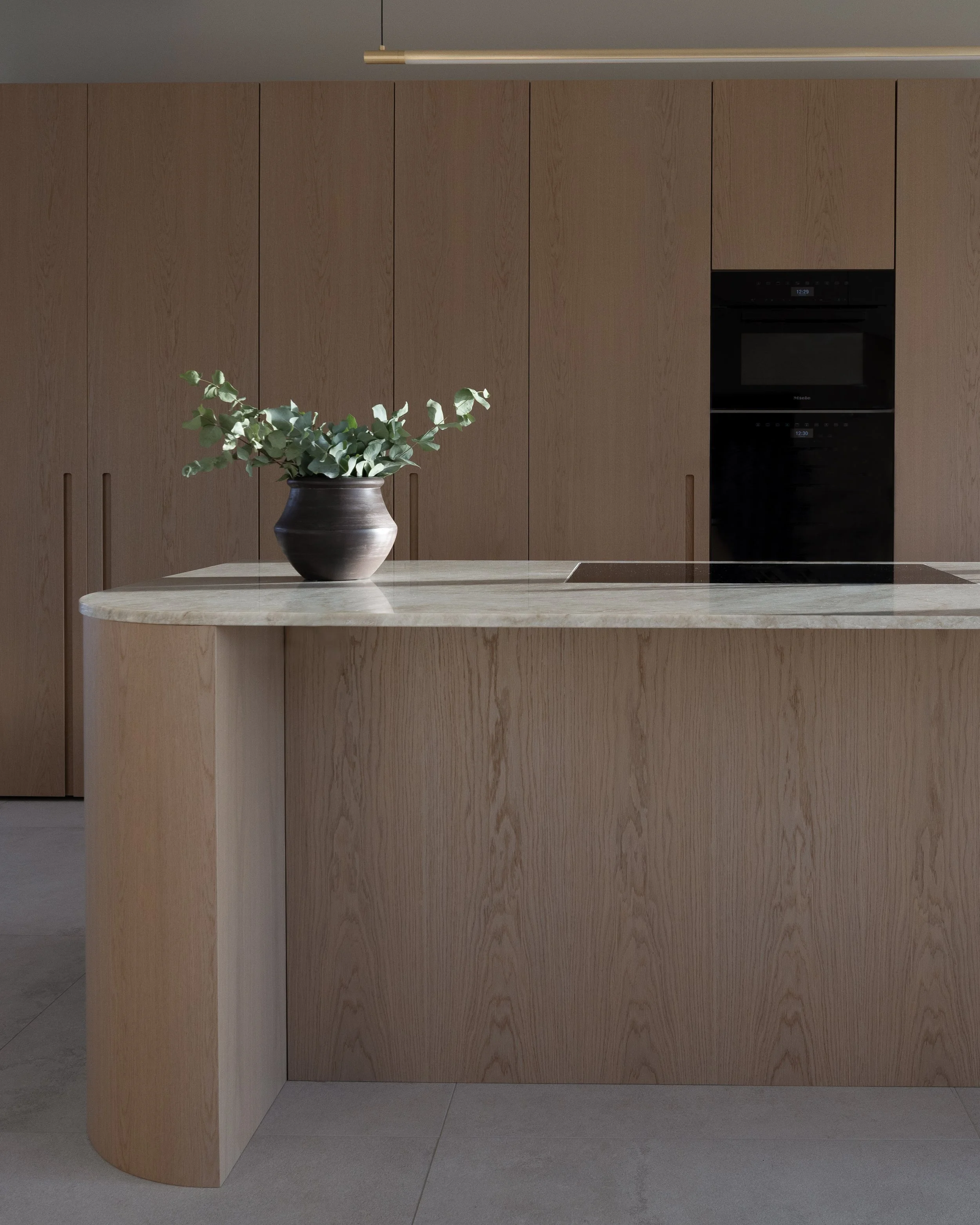 A modern kitchen with wood cabinetry and a marble countertop island, featuring a black vase with eucalyptus leaves, and built-in black appliances.