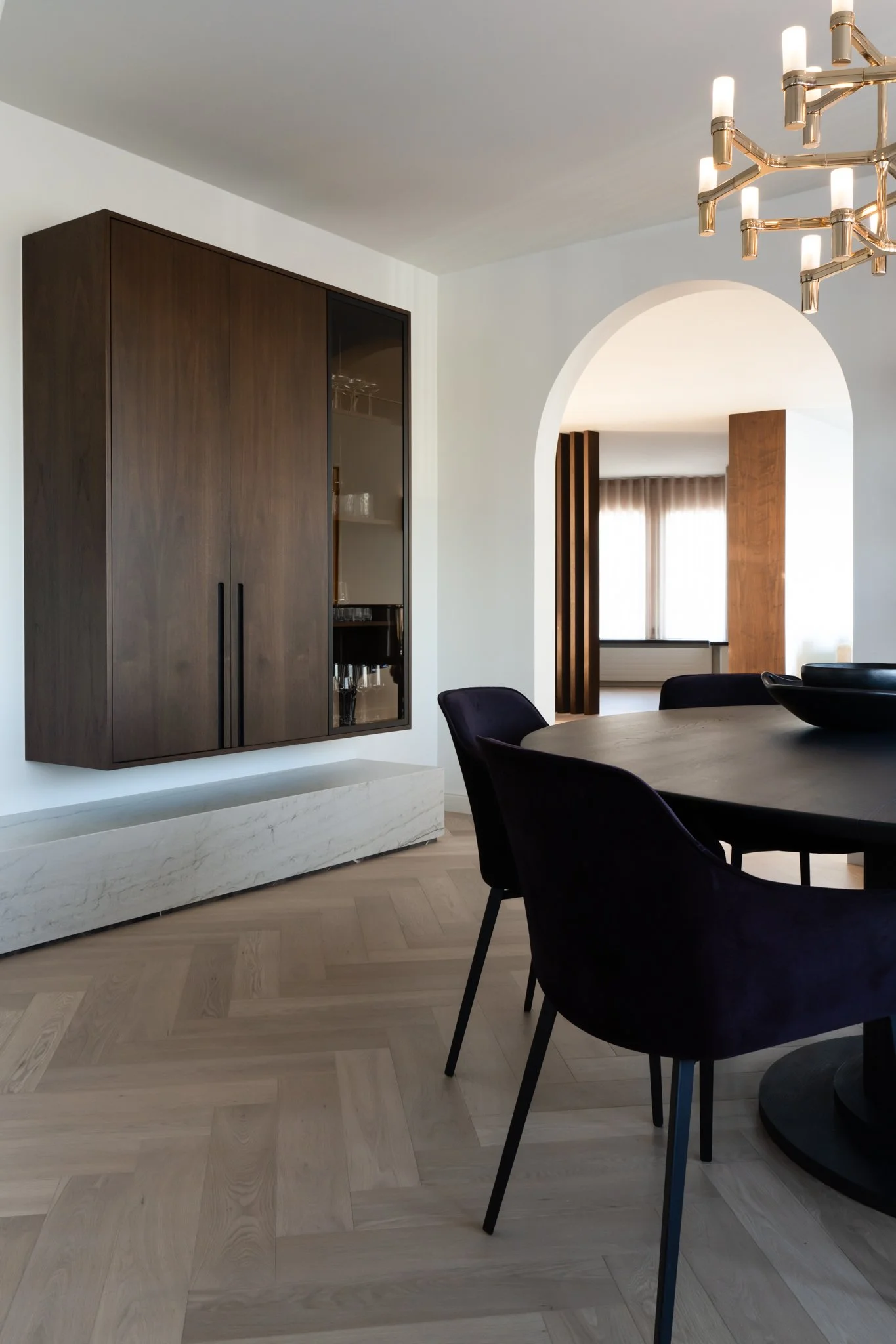 A modern dining room with a round table, dark upholstered chairs, a wooden cabinet with glass doors, an arched doorway, and a contemporary chandelier.