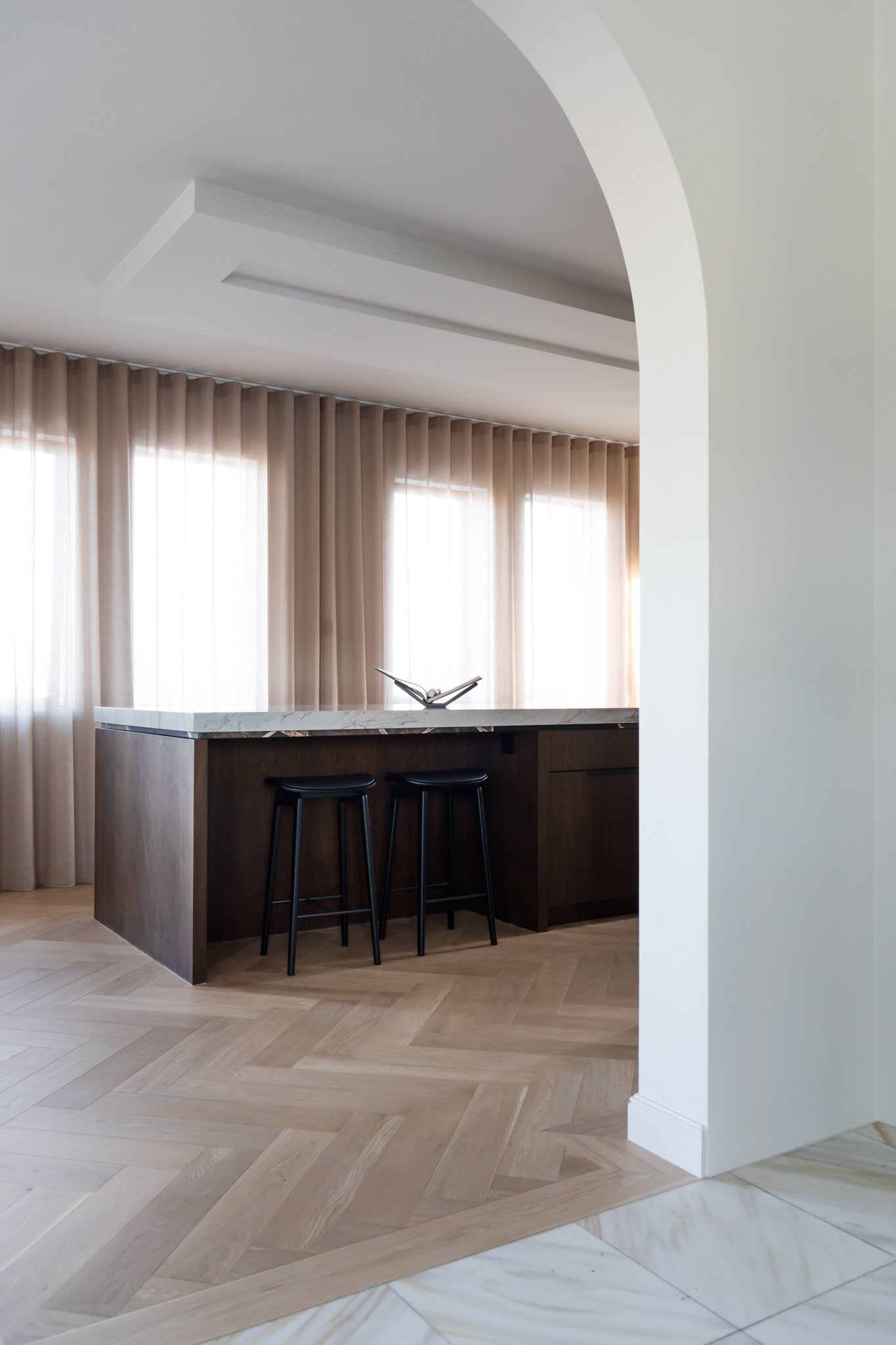 A kitchen island with a marble countertop and three black bar stools, set in front of beige curtains and large windows with natural light.