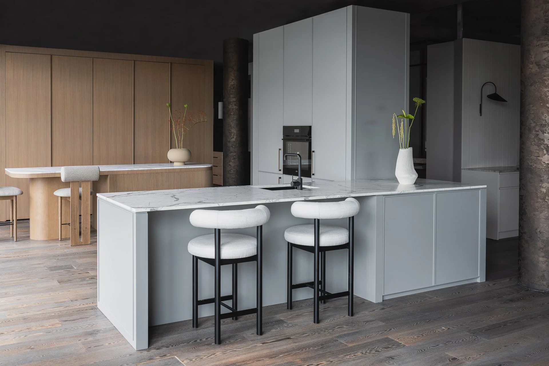 Modern kitchen with white marble island, black bar stools, light wood cabinetry, and potted plants.