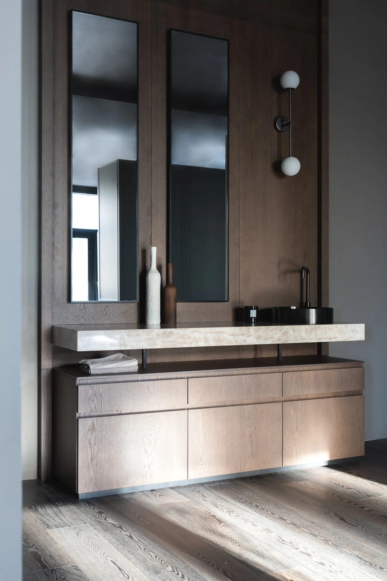Modern bathroom vanity with two large mirrors, a black sink with a gold faucet, and wooden cabinets below. Decorative vases are placed on the counter, with a wall-mounted light fixture above.