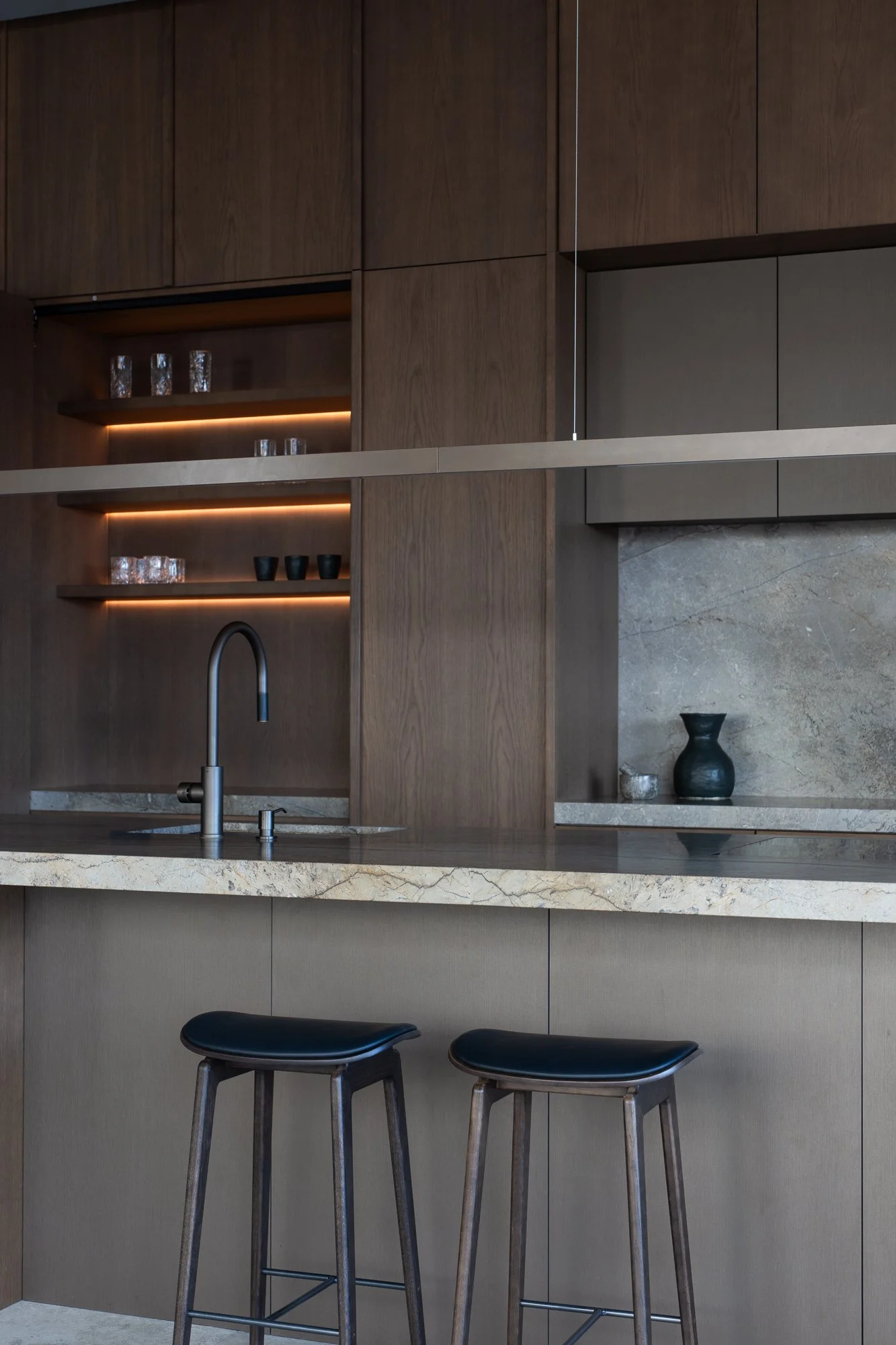 Modern kitchen with wooden cabinets, open shelves with glasses and cups, a marble countertop, a sink with a black faucet, and two black bar stools in front of a beige wall.