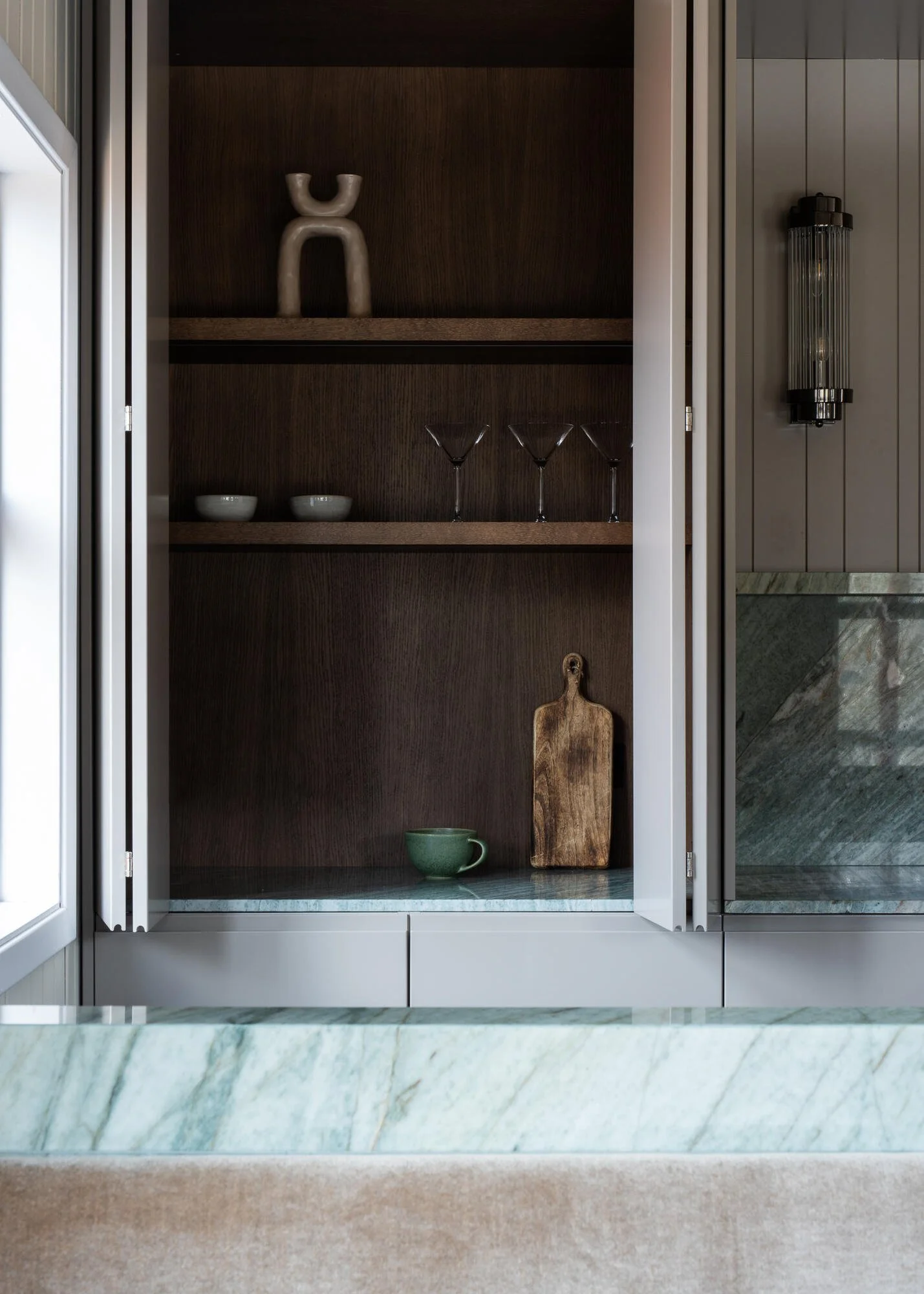 Open kitchen cabinet with dark wood interior holding decorative items, bowls, martini glasses, a cutting board, and a green mug, with marble countertop in foreground.