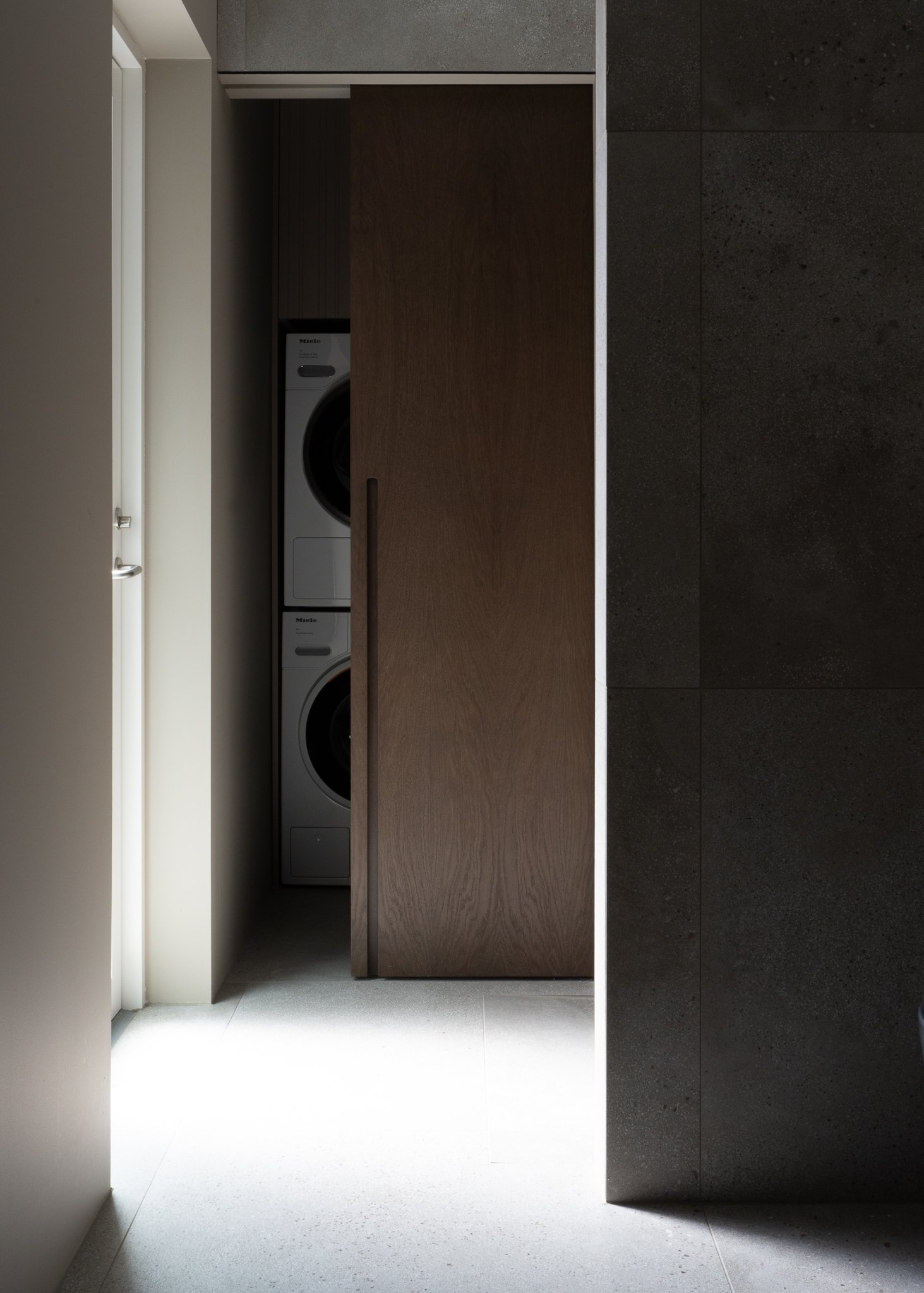 Modern laundry room with a white washer and dryer stacked behind a sliding wooden door, gray dark tiled floor, and white walls illuminated by natural light from a window.