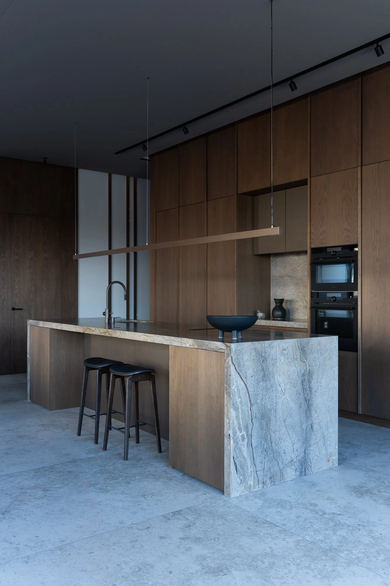 Modern kitchen with a large marble island, dark wood cabinets, and black bar stools, featuring minimalist decor.