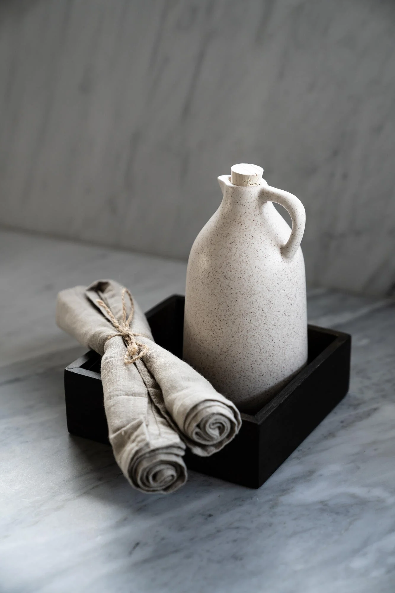 A white ceramic pitcher with a handle and a wooden stopper, placed in a black tray on a marble surface; next to it are two rolled cloth napkins tied with twine.