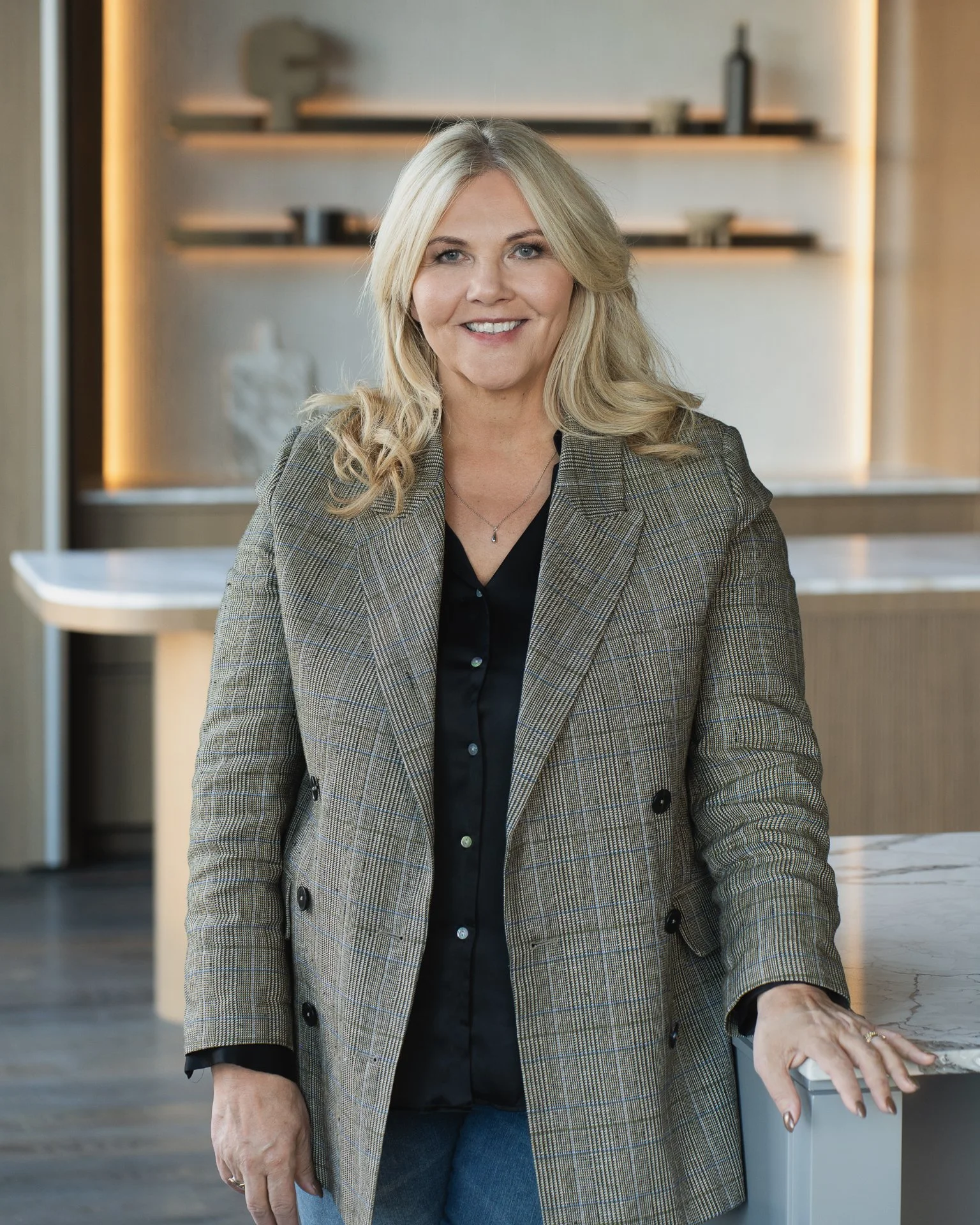 A woman with shoulder-length blonde hair smiling, wearing a plaid blazer over a black shirt, standing in a modern kitchen with a marble countertop and wooden shelves with minimal decor in the background.