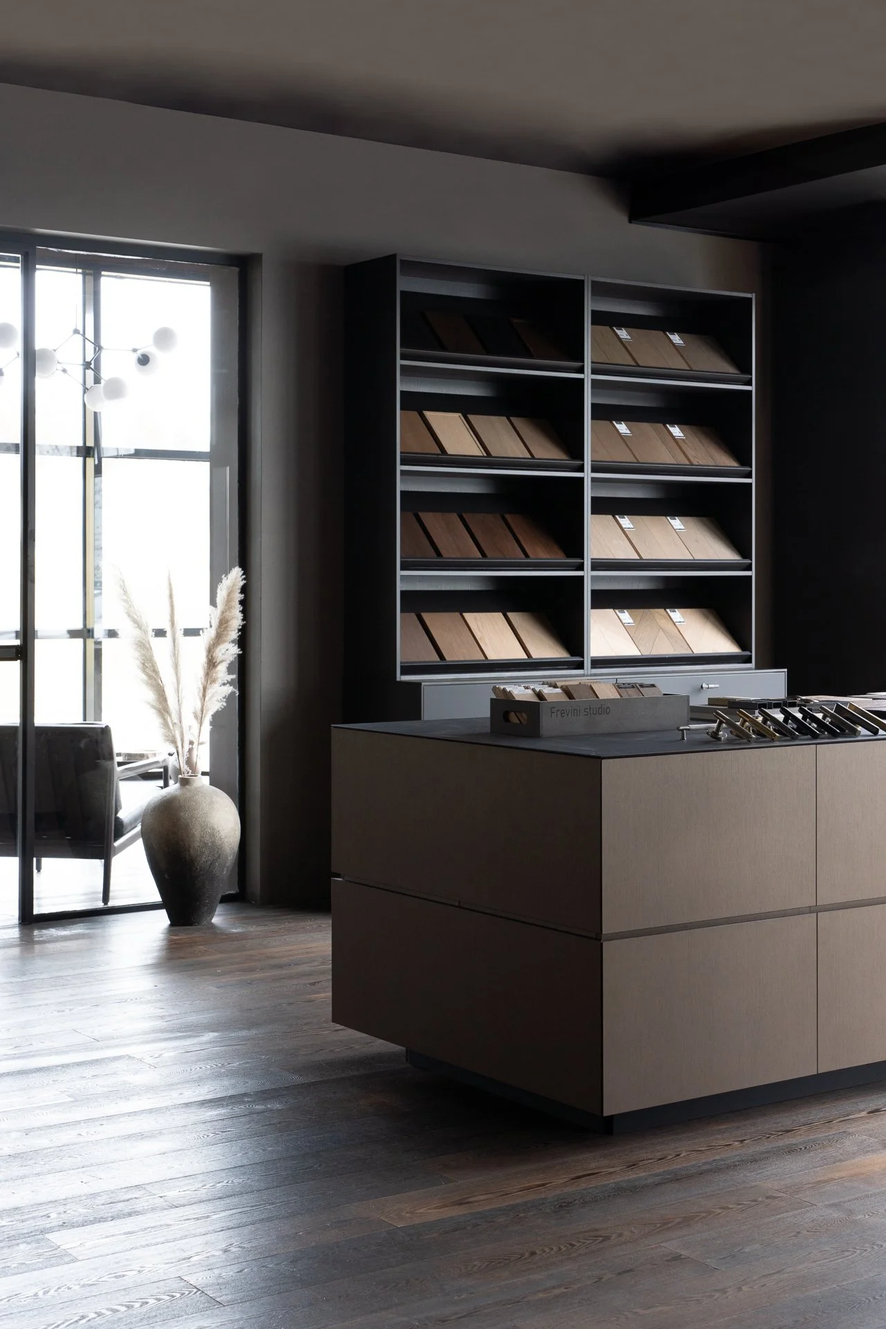 Modern interior space featuring a display of wooden flooring samples, a large beige kitchen island, a window with natural light, and a decorative vase with pampas grass.