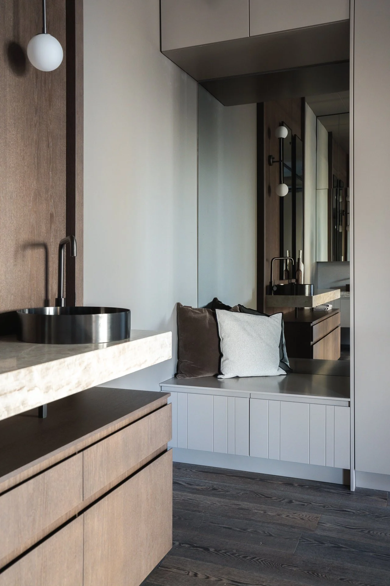 Modern kitchen with floating beige marble countertop, black sink, wooden cabinets, black fixtures, and a window seat with pillows, dark hardwood floors, and a mirror.