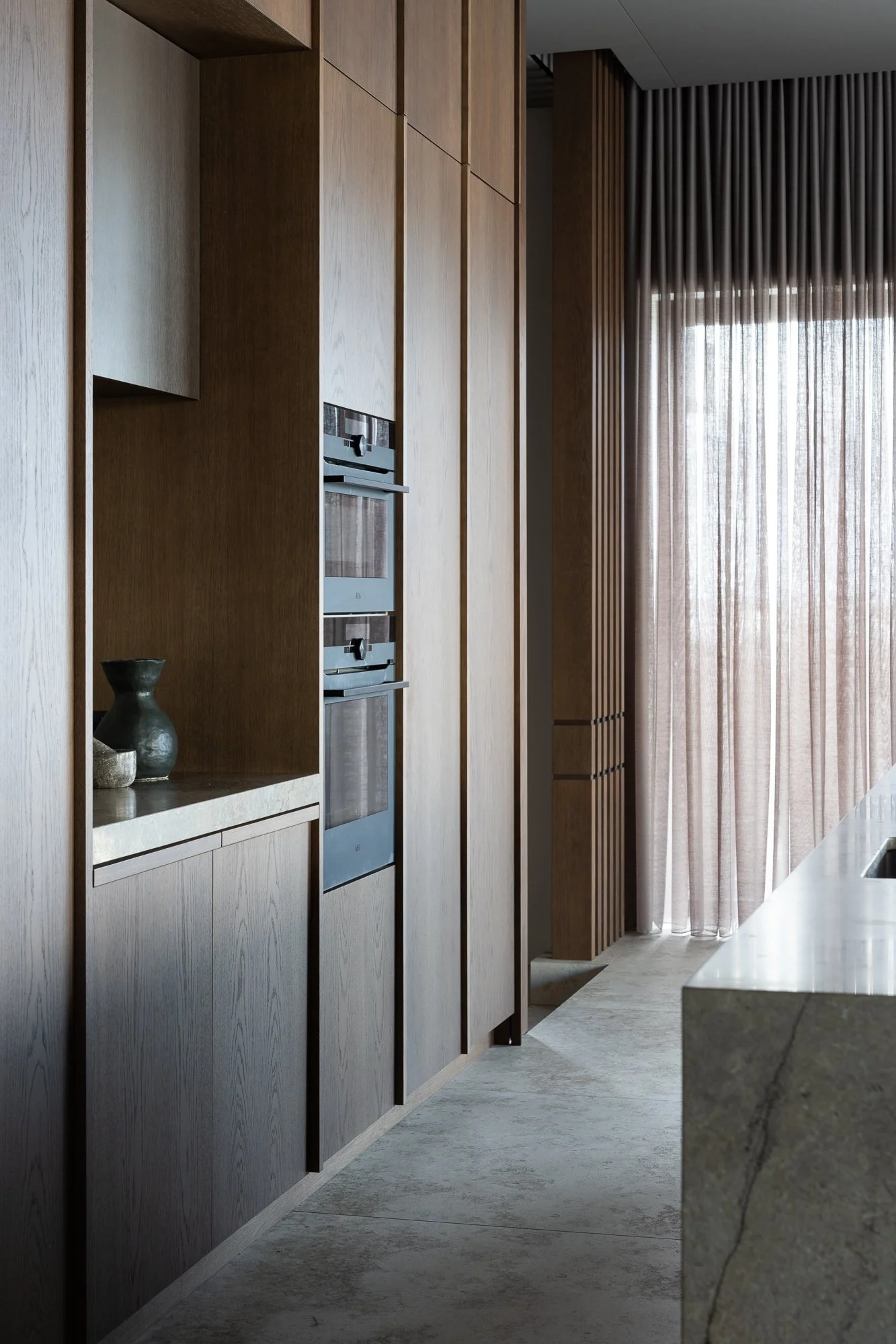 Modern kitchen with wooden cabinets, built-in stainless steel oven, and sheer curtains covering a large window.