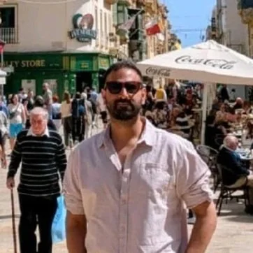 Man with sunglasses and beard standing outdoors in a busy street with shops and outdoor cafe seating.