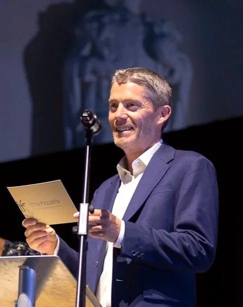 A man in a navy suit and white shirt standing at a podium, smiling and holding a paper, with a microphone in front of him. There is a stage backdrop behind him.