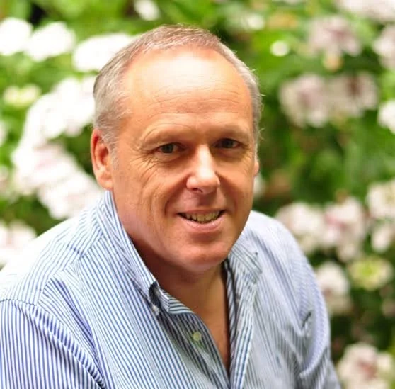 Portrait of a middle-aged man with short, light-colored hair, smiling, wearing a light blue striped button-up shirt, outdoors with blurred greenery and flowers in the background.