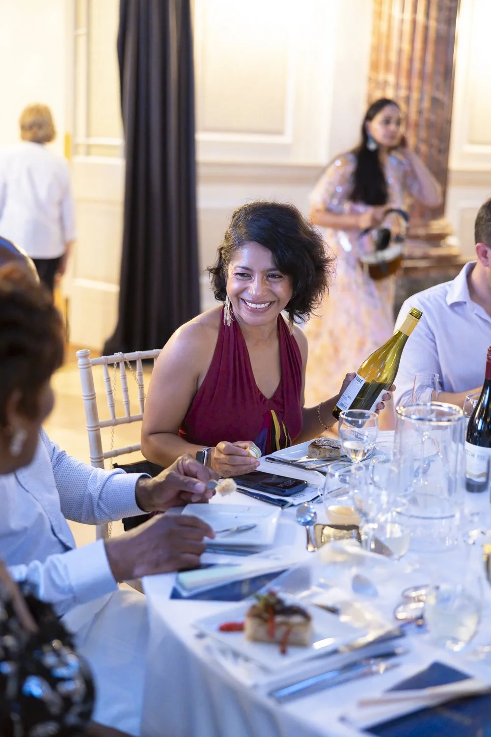 A woman in a red dress smiling at a dinner table with wine and food, holding a wine bottle, during a social event or celebration.