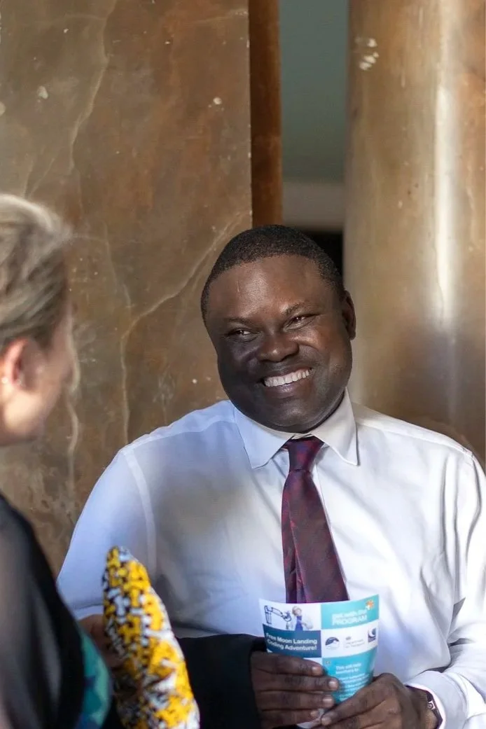 Smiling man in white shirt and maroon tie talking to woman at an indoor event.