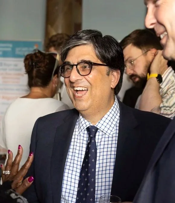 A man wearing glasses, a navy suit, and a checkered shirt, smiling and engaging in conversation at a social event.