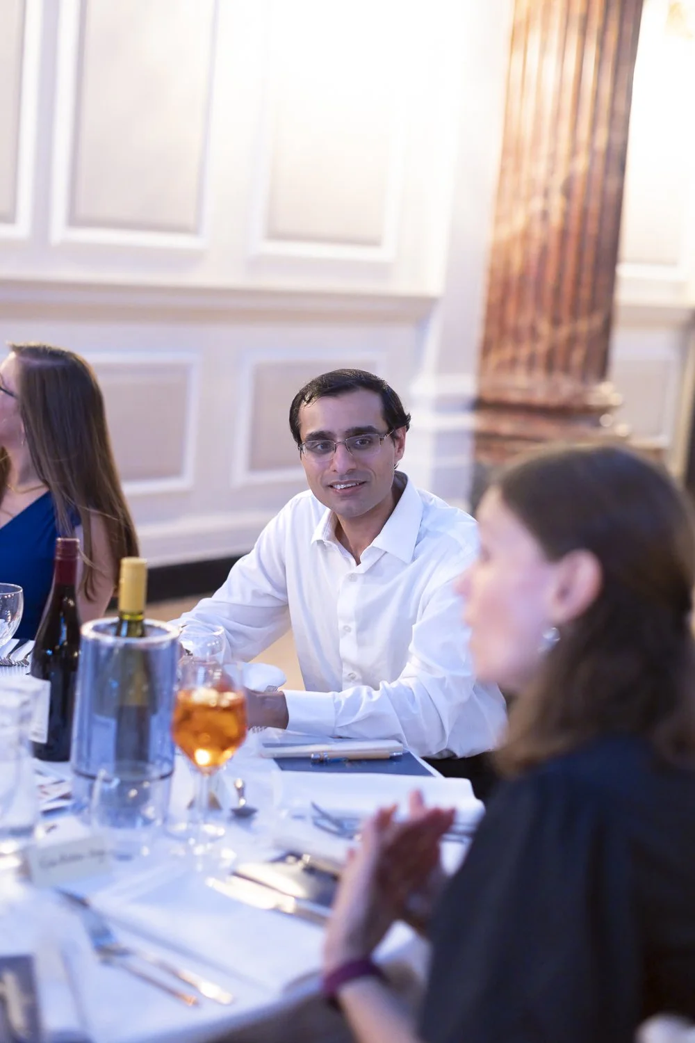 Group of people dining at a formal event, with a man in a white shirt smiling and looking at the camera.
