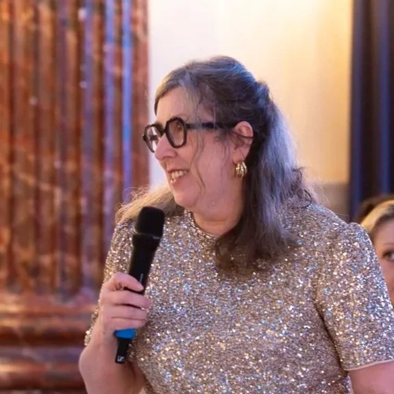 A woman with long hair, glasses, and gold earrings holding a microphone and smiling during an event indoors.