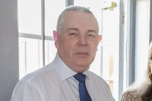 A middle-aged man with short gray hair wearing a white dress shirt and a dark blue tie, standing indoors near a window.