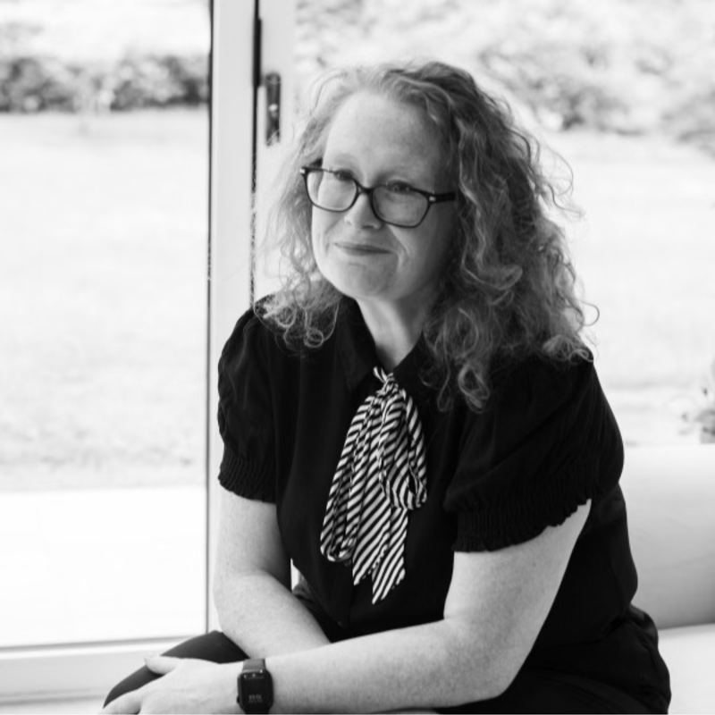A woman with curly hair and glasses sitting by a window, wearing a black blouse with a striped bow tie, with a slight smile.