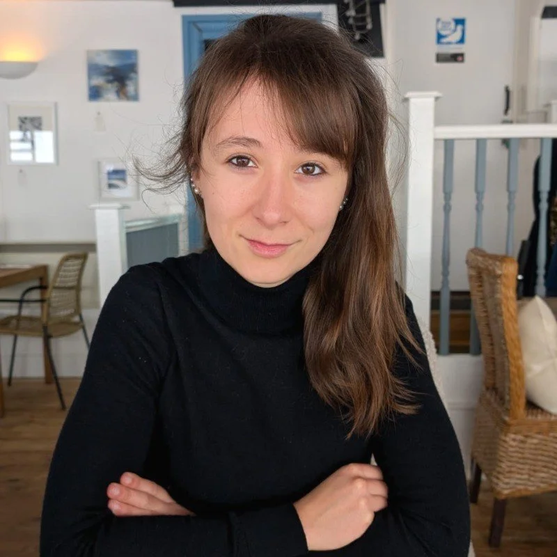 A woman with long brown hair and a black turtleneck sitting indoors with her arms crossed, smiling subtly at the camera.
