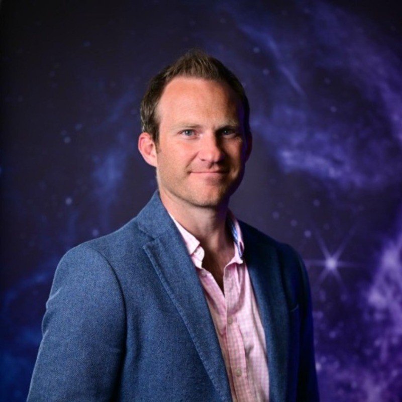 A man in a blue blazer and pink shirt standing against a galaxy-themed background.